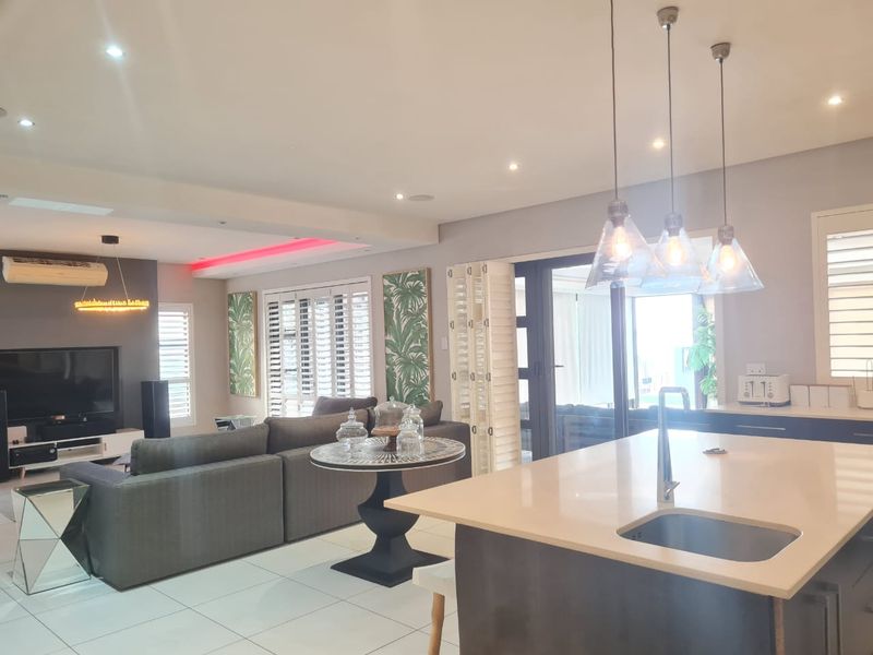 Spacious open-plan living area featuring a modern kitchen island with sink, pendant lighting, tiled floors, and plantation shutters leading to an outdoor space.