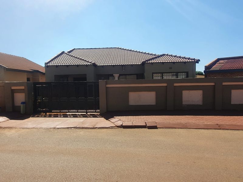 Front view of a single-story house featuring a tiled roof, secure perimeter wall with a metal gate, and paved street-side access.