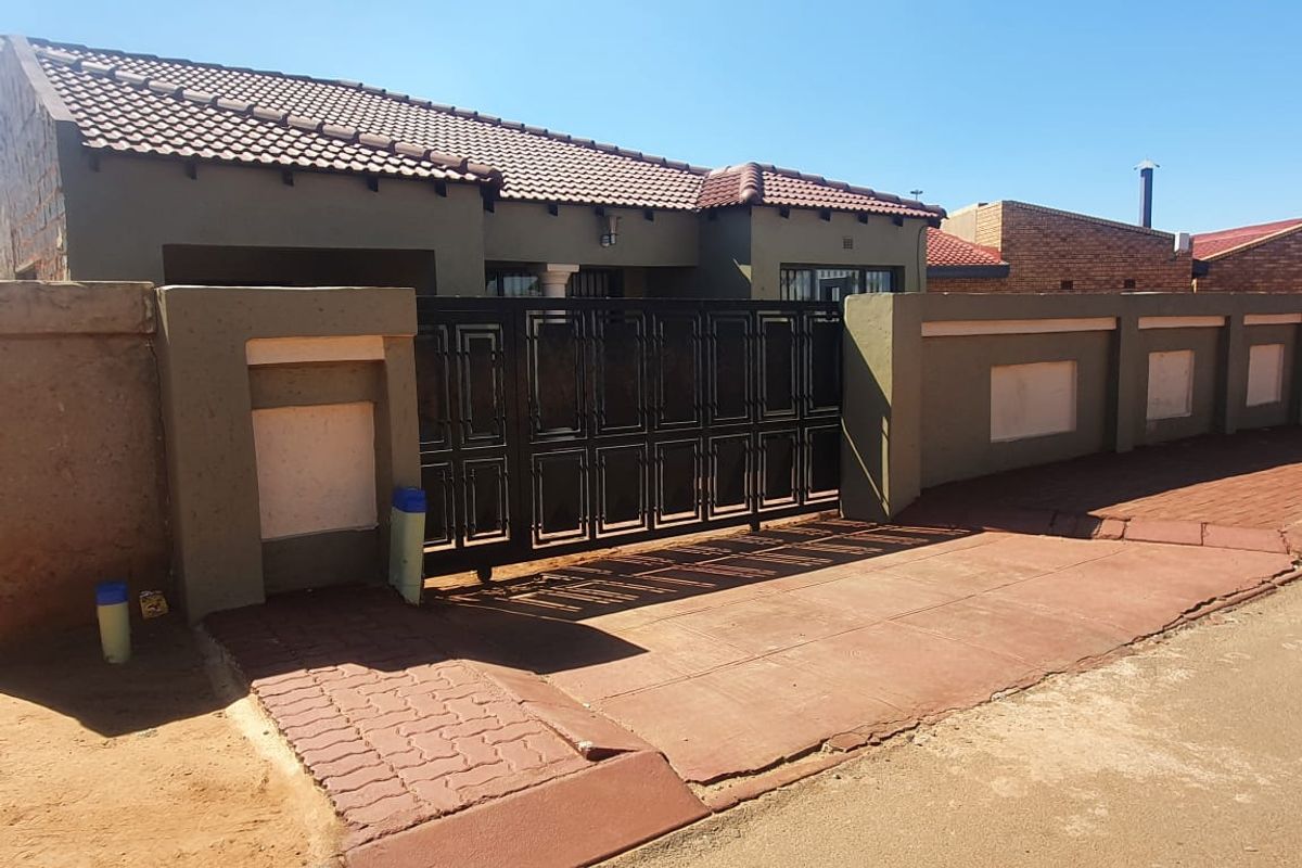 Front exterior view of the property featuring a tiled roof, secure metal driveway gate, and a paved entrance area with perimeter boundary walls.