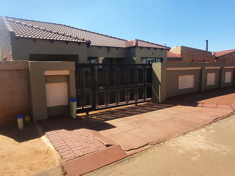Front exterior view of the property featuring a tiled roof, secure metal driveway gate, and a paved entrance area with perimeter boundary walls.