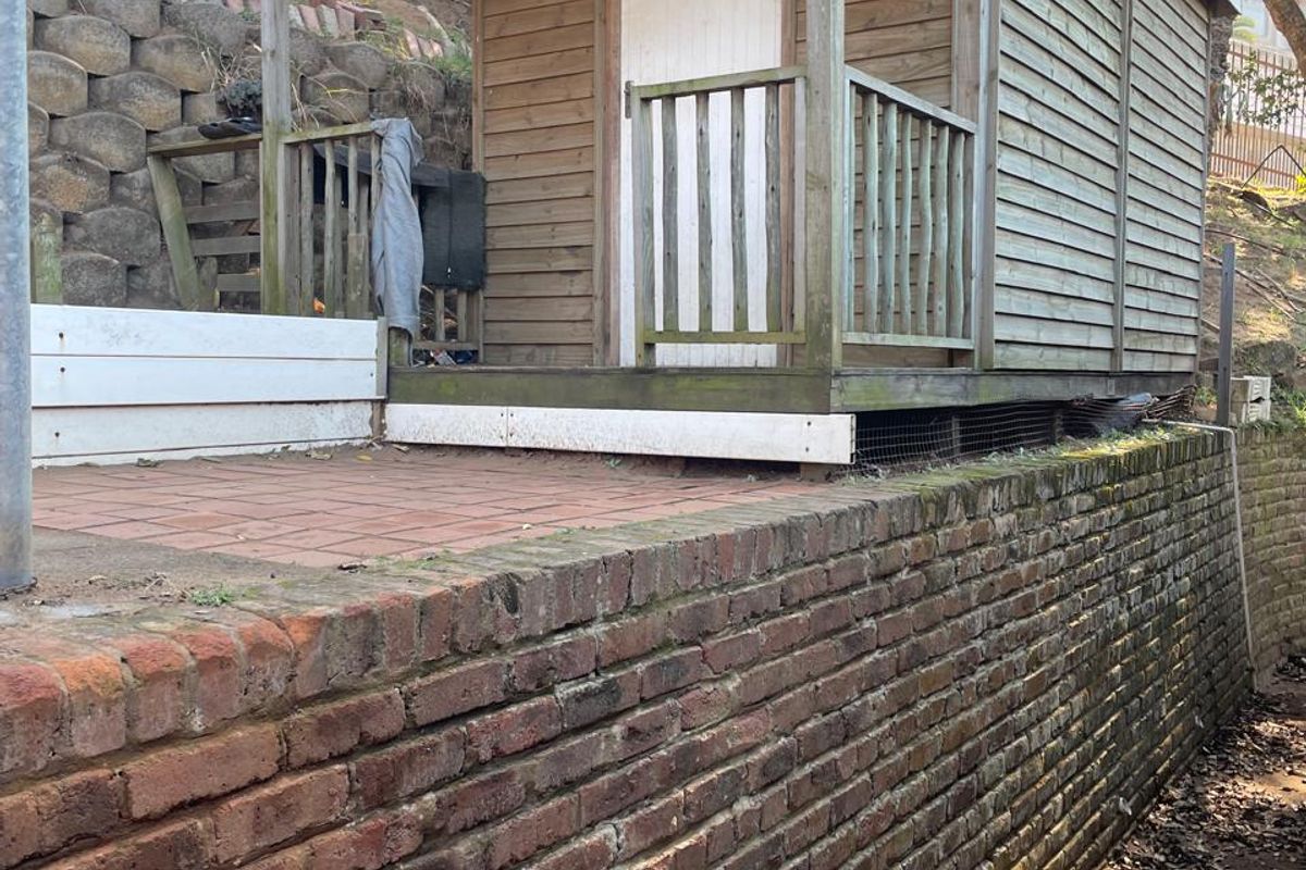 A detached timber shed structure situated on a raised paved patio area, supported by a substantial brick retaining wall in the rear garden.