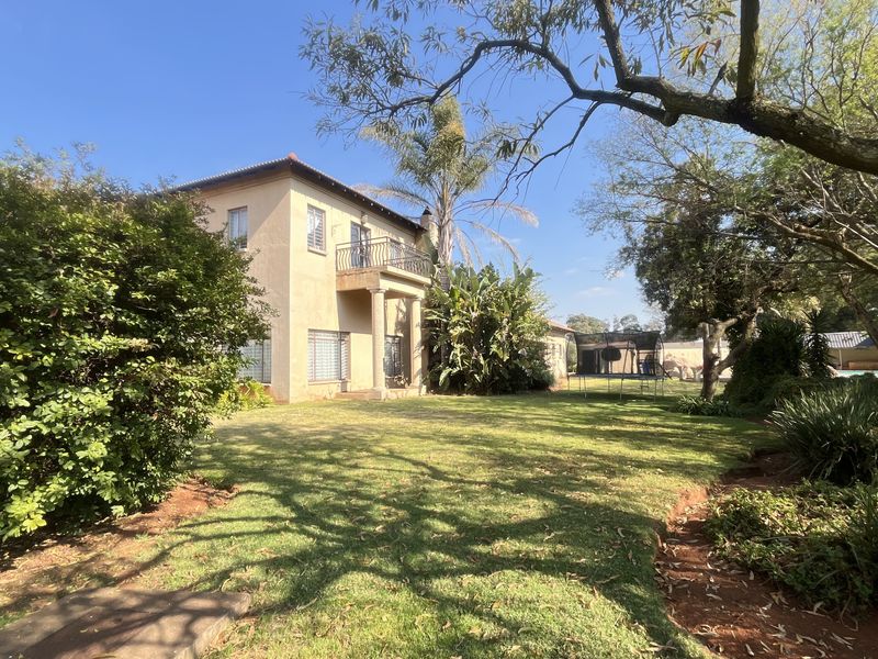 Rear view of a double-story residence featuring a balcony with columns, set within a spacious, established garden with mature trees.