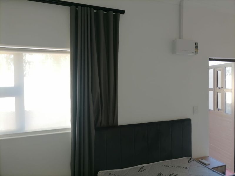 A clean bedroom featuring a window with dark curtains, a wall-mounted air conditioning unit, and a doorway leading to an adjacent space.