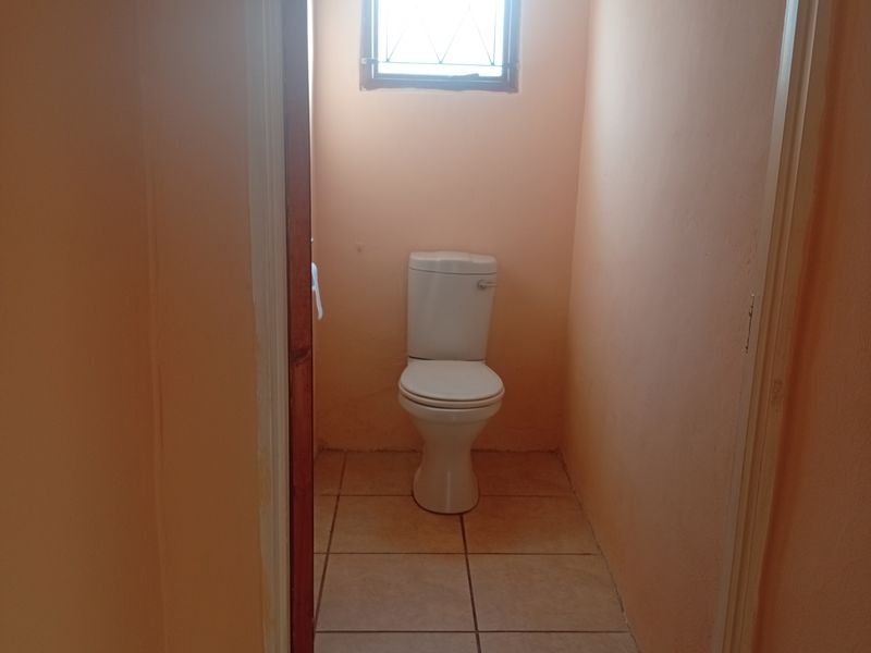 A small, functional guest toilet featuring tiled flooring, a window with security bars, and a ventilation grille.