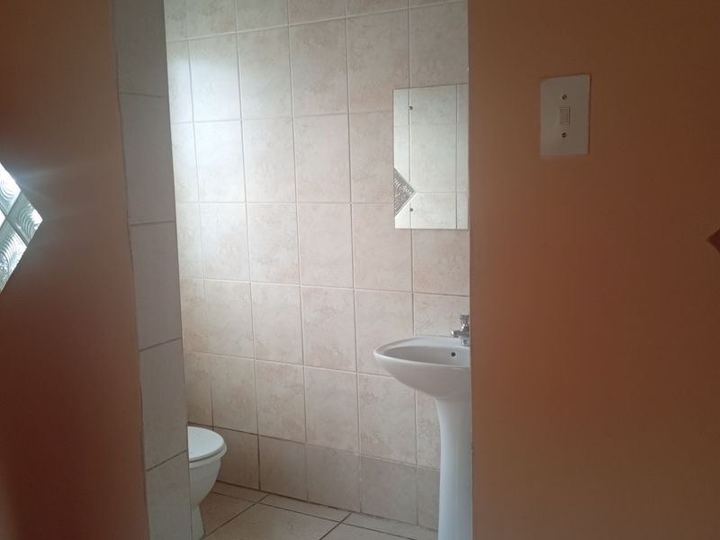 A functional bathroom featuring a pedestal sink, toilet, and neutral wall and floor tiling, viewed through a doorway.
