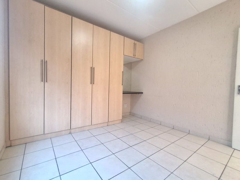 A clean, empty bedroom featuring light wood-finish built-in cupboards, a matching built-in desk, and neutral tiled flooring.