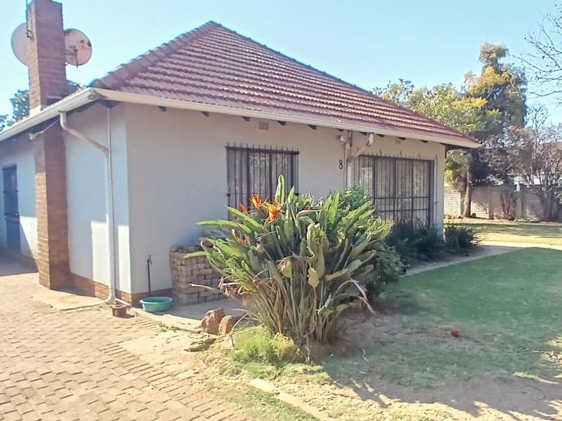 Front view of a single-story house featuring a tiled roof, brick chimney, paved driveway, carport, and a front garden with security bars on windows.