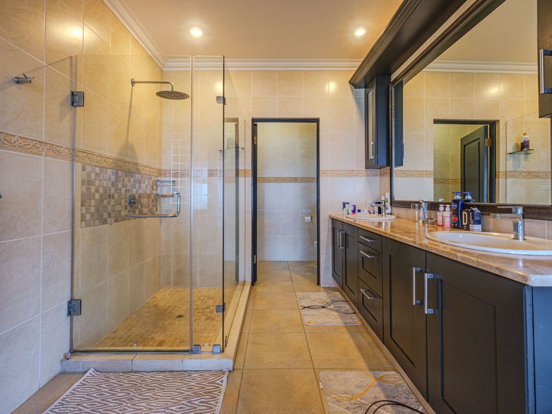A well-maintained bathroom featuring a glass-enclosed rain shower, a double vanity with stone countertops, and neutral wall tiling.