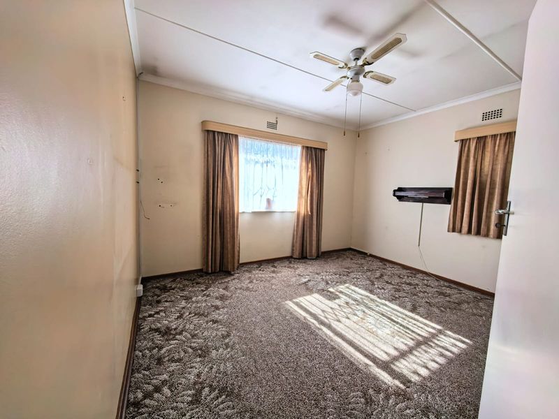 A spacious, empty bedroom featuring carpeted flooring, a ceiling fan, and dual windows with existing curtain rails.