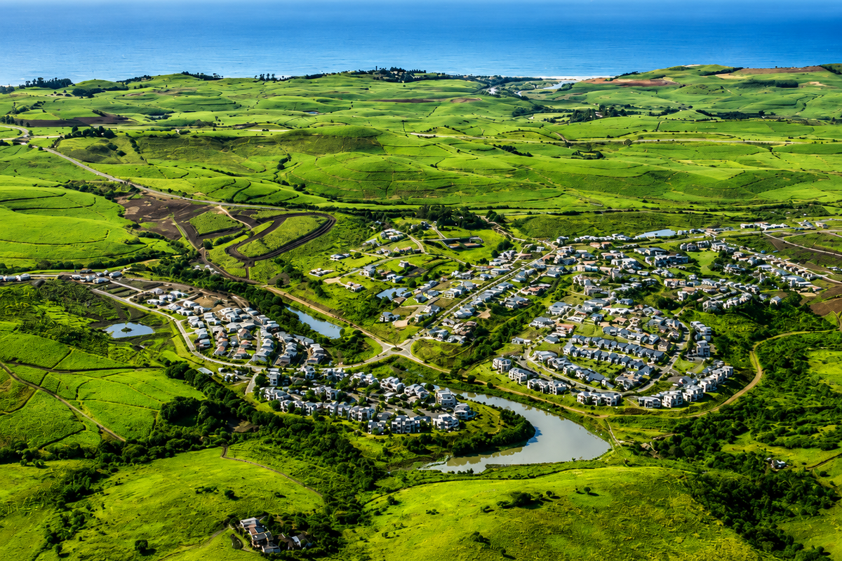 An expansive aerial view of a residential estate nestled within lush green rolling hills, featuring a central lake and proximity to the coastline.