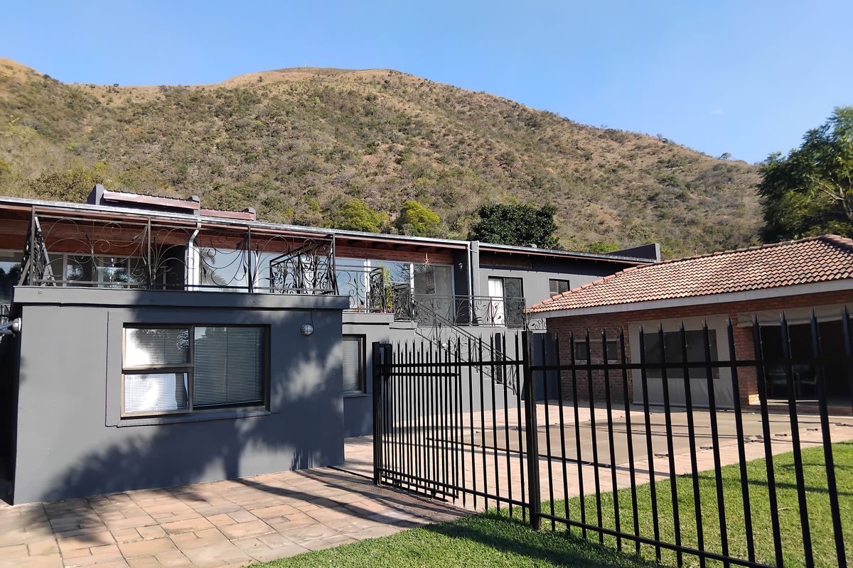 Rear exterior view of the property featuring a paved patio, balcony with wrought iron railings, and a swimming pool area set against a mountain backdrop.