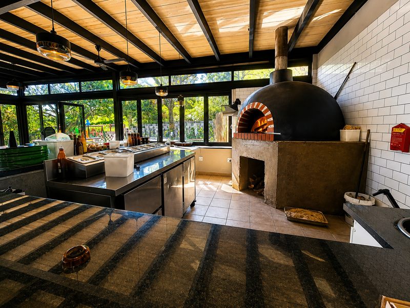 A professional-grade outdoor kitchen area featuring a large wood-fired pizza oven, stainless steel prep stations, and expansive windows overlooking the garden.