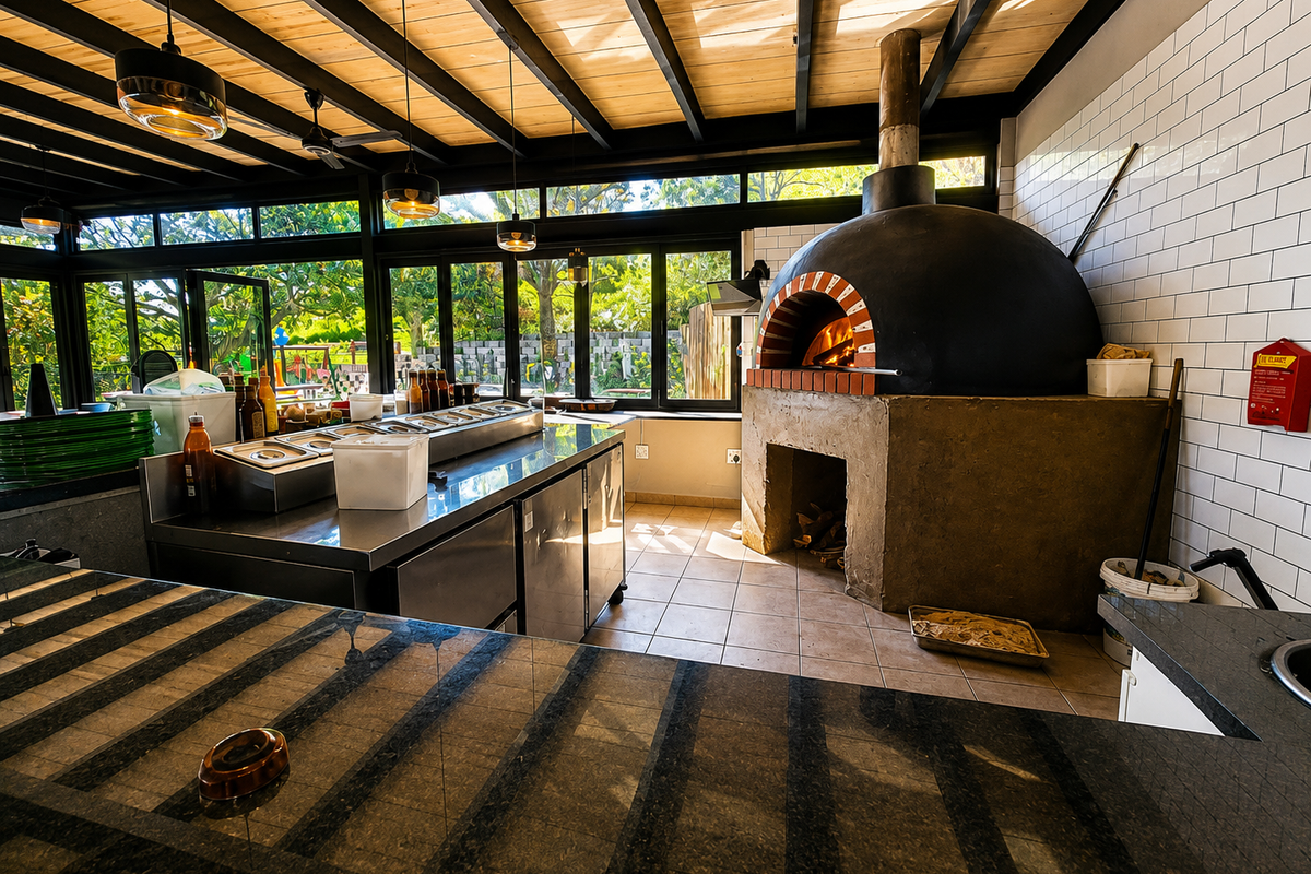 A professional-grade outdoor kitchen area featuring a large wood-fired pizza oven, stainless steel prep stations, and expansive windows overlooking the garden.