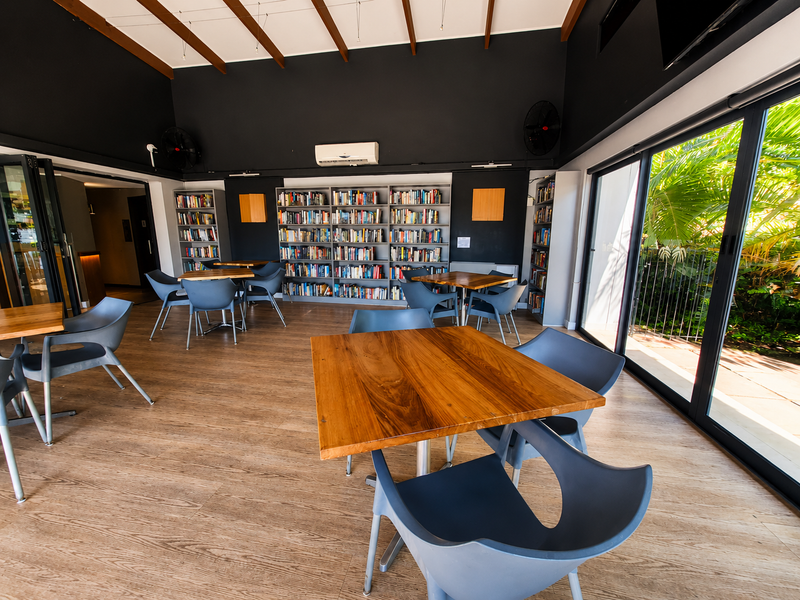 A modern, well-lit communal living space featuring built-in bookshelves, exposed timber ceiling beams, and large sliding glass doors opening to the garden.