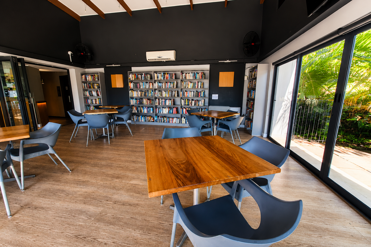 A modern, well-lit communal living space featuring built-in bookshelves, exposed timber ceiling beams, and large sliding glass doors opening to the garden.