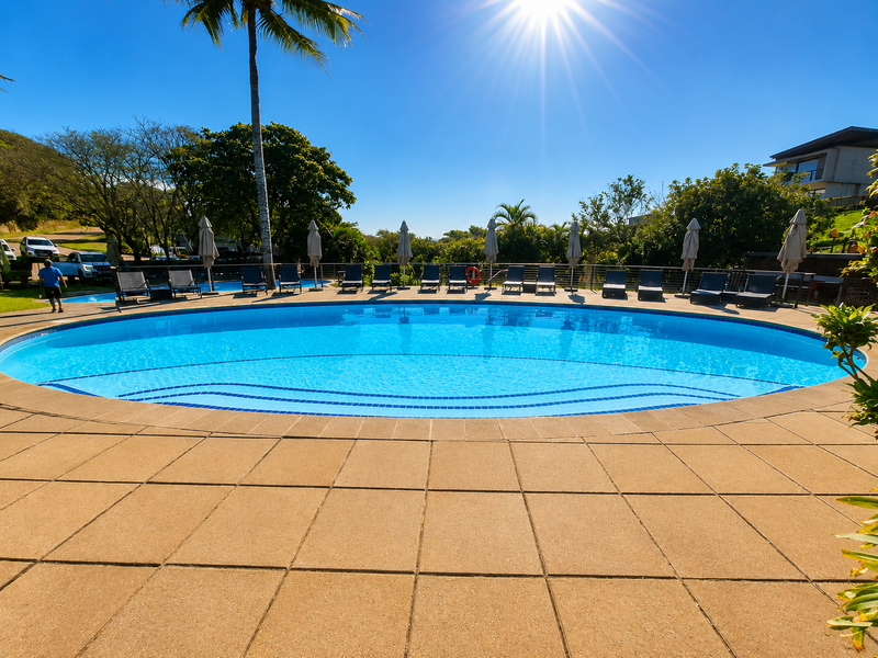 A well-maintained circular swimming pool with a paved deck area, surrounded by lush landscaping and metal safety fencing.
