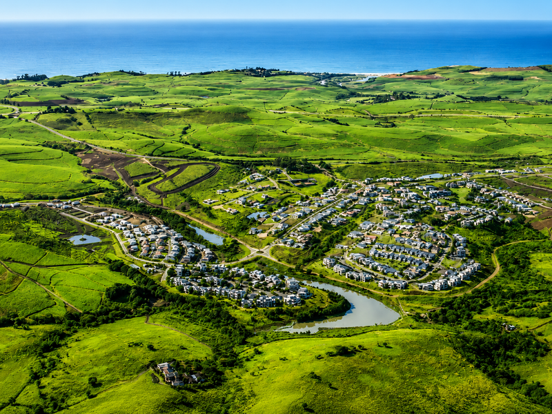 An expansive aerial view of a residential estate nestled within lush green rolling hills, featuring a central lake and proximity to the coastline.