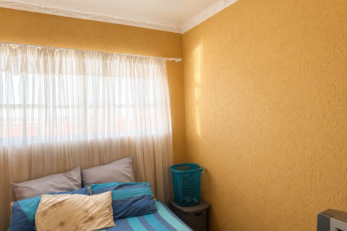 A bedroom featuring textured yellow walls and decorative ceiling cornicing, providing a functional space with natural light.