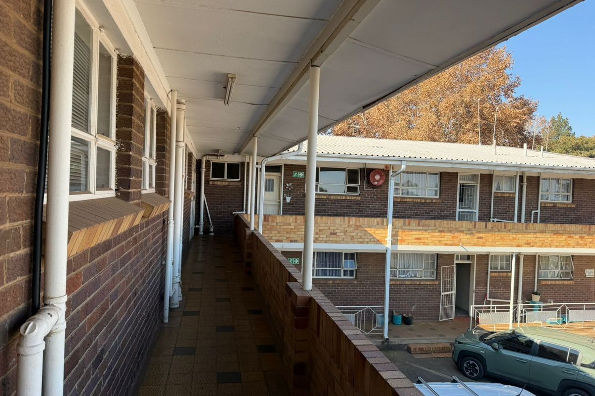 An exterior view of a multi-story flat complex featuring a covered walkway, brick facade, and tiled balcony area overlooking the parking space.