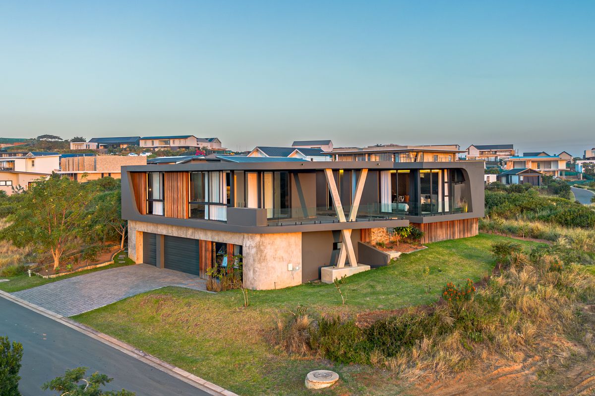 Aerial view of a modern architectural home featuring timber cladding, concrete structural elements, glass balustrades, and a double garage in a landscaped setting.