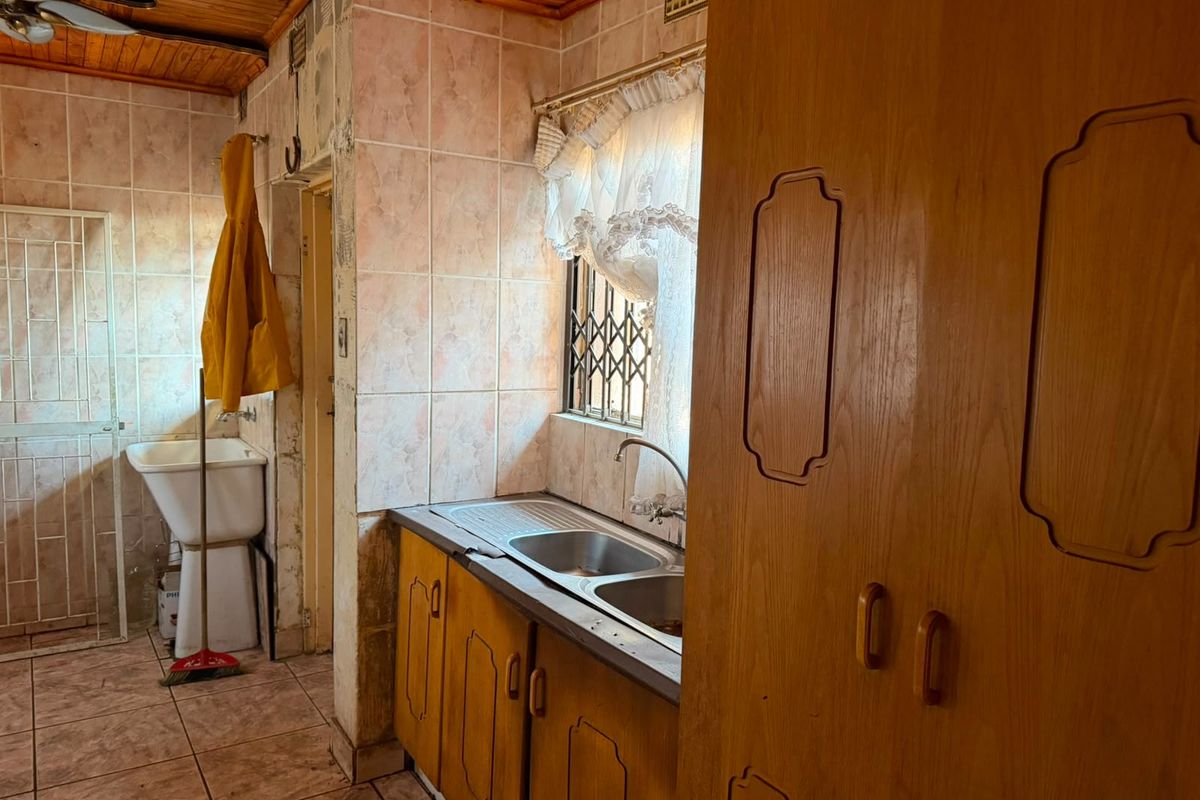 A dated kitchen featuring wooden cabinetry, tiled walls, and a double stainless steel sink, with a separate utility sink area and timber ceiling.