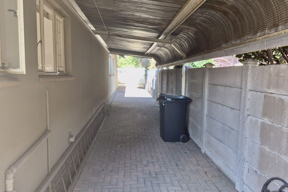 A narrow side passage featuring a paved walkway, covered by a corrugated metal roof structure and enclosed by precast concrete boundary walls.