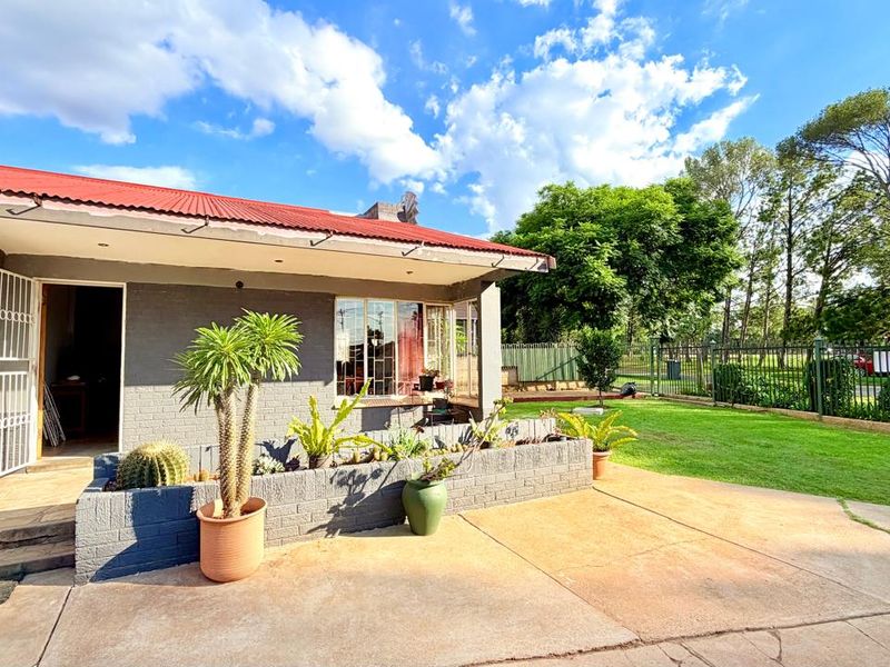 Rear exterior view of the house featuring a concrete patio, brick planter box, and a corrugated metal roof, set against a landscaped garden.