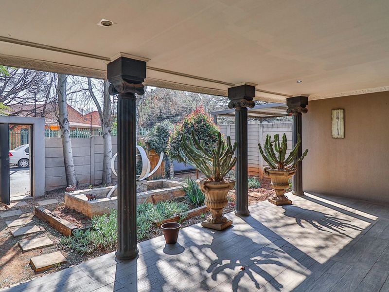 A covered patio area featuring decorative columns, tiled flooring, and recessed lighting, overlooking a landscaped garden with a built-in water feature.