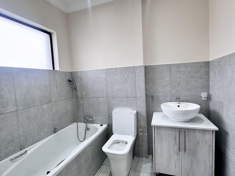 Modern bathroom featuring a bathtub with handheld shower, a sleek vanity with vessel sink, and neutral grey floor-to-ceiling tiling.