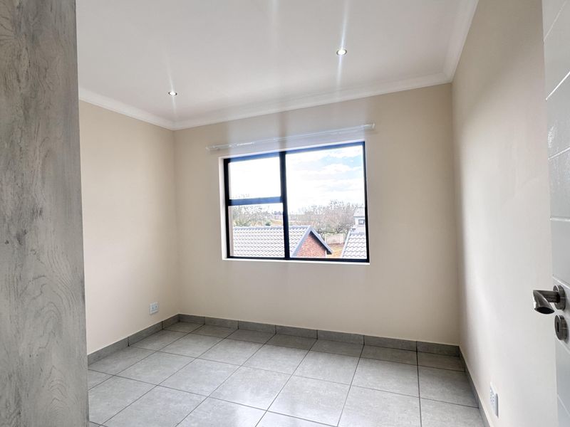 A bright, empty bedroom featuring neutral walls, modern grey tiled flooring, and recessed downlighting with a view through an aluminium-framed window.