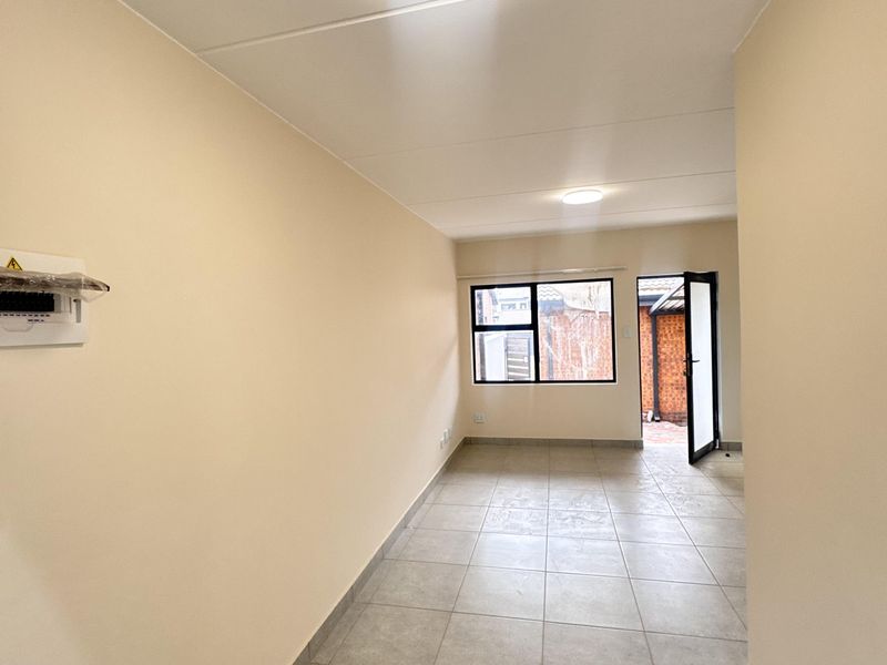 A bright, empty living area featuring neutral tiled flooring, flush mount lighting, and direct access to an outdoor space through a glass-paneled door.