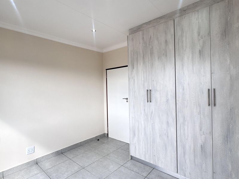A clean, empty bedroom featuring modern grey wood-look built-in cupboards, neutral tiled flooring, and recessed ceiling downlights.