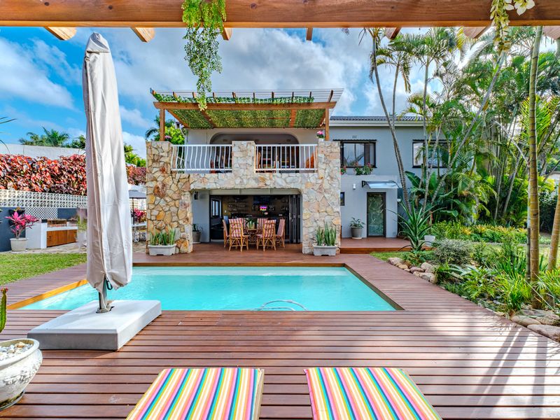 A beautifully landscaped outdoor entertainment area featuring a swimming pool, timber decking, stone-clad structure, and a covered patio with built-in braai facilities.