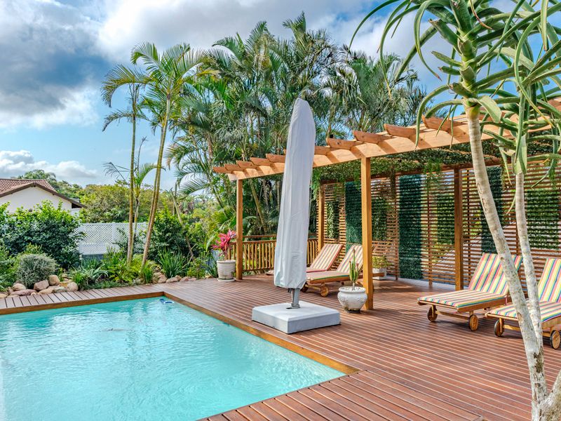 A well-maintained outdoor pool area featuring timber decking, a wooden pergola, and a privacy screen, set within a landscaped garden environment.