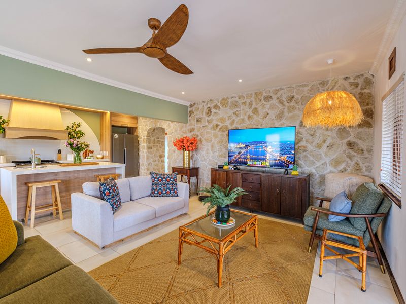 A bright open-plan living and kitchen area featuring a striking stone feature wall, modern kitchen island, and a wooden ceiling fan.