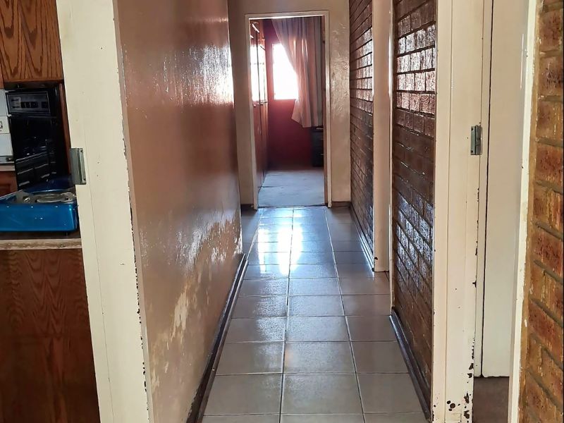 A narrow hallway featuring tiled flooring, exposed brickwork, and a timber ceiling, showing signs of wear and moisture damage on the painted wall.