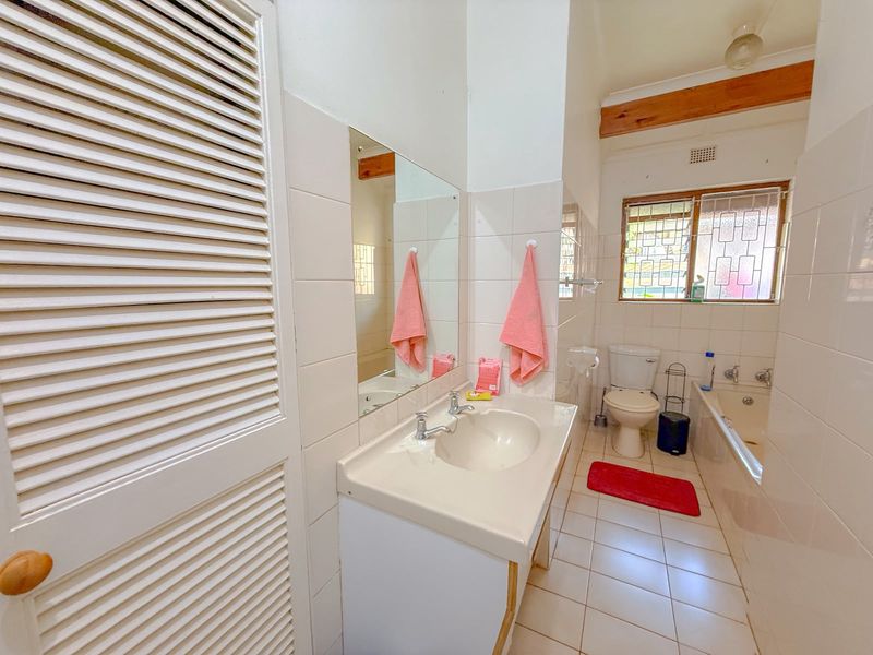A functional bathroom featuring a vanity basin, built-in bathtub, toilet, and louvered storage cupboard, finished with neutral wall and floor tiles.