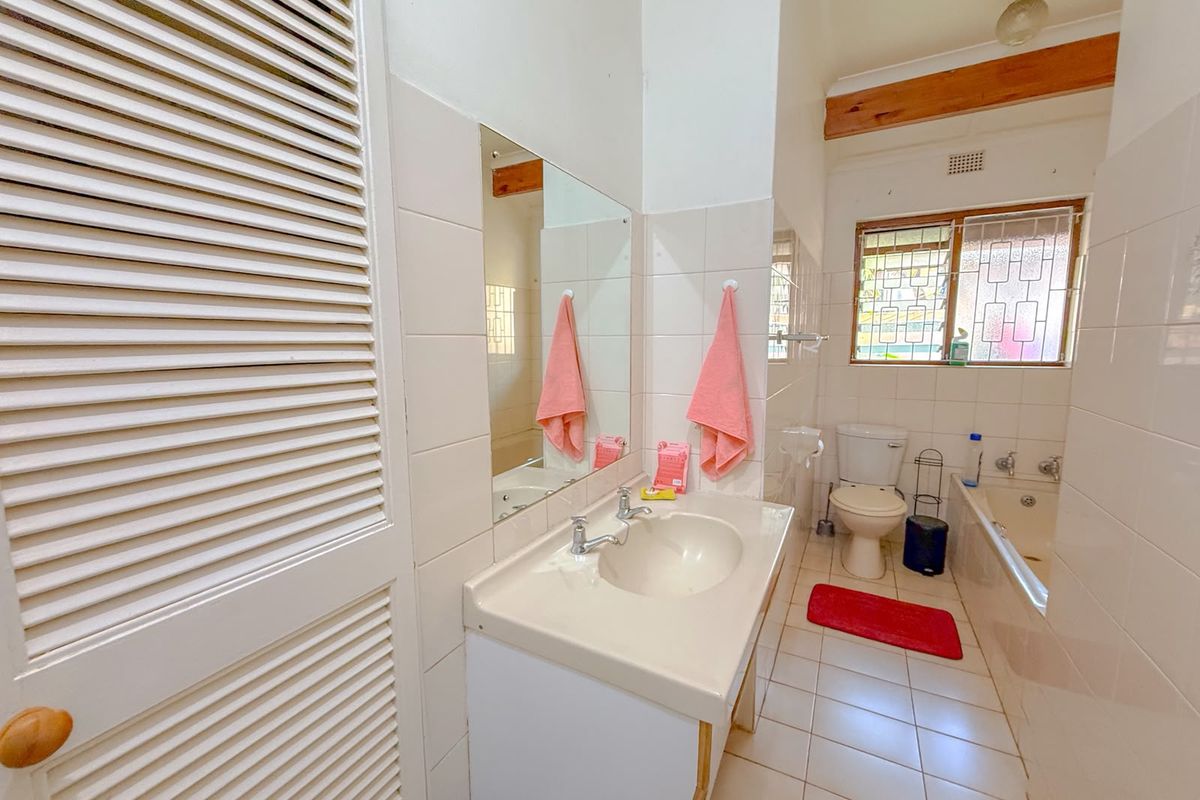 A functional bathroom featuring a vanity basin, built-in bathtub, toilet, and louvered storage cupboard, finished with neutral wall and floor tiles.