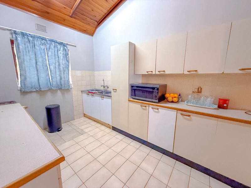 A functional kitchen featuring built-in cabinetry, laminate countertops, a stainless steel sink, and exposed timber ceiling beams with tiled flooring throughout.