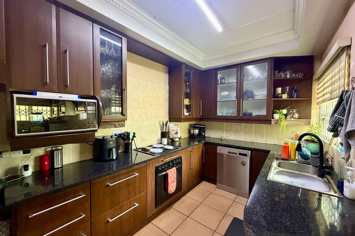 A functional kitchen featuring dark wood cabinetry, granite countertops, built-in oven, hob, and integrated dishwasher with tiled flooring and decorative ceiling detailing.