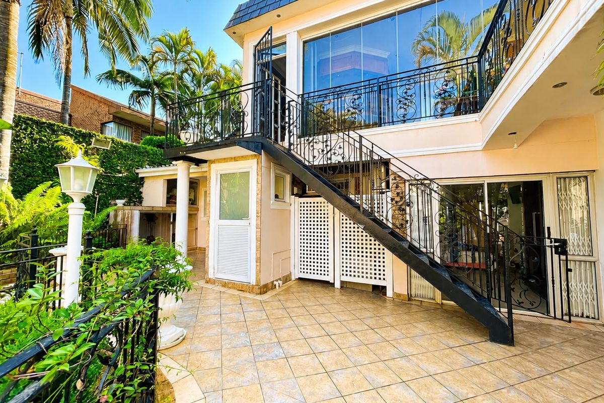 Rear exterior view featuring a tiled patio, a wrought iron staircase leading to an enclosed upper balcony, and established garden surroundings.