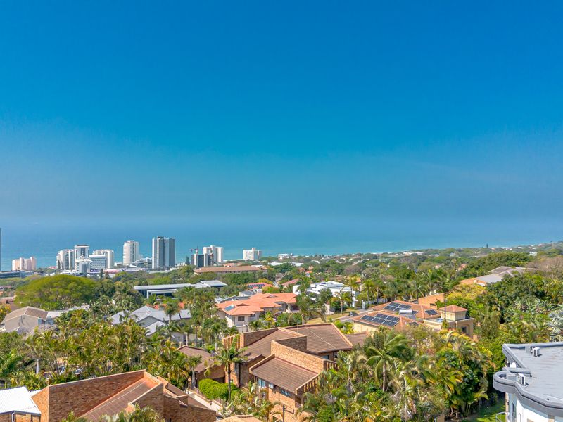 Elevated panoramic view overlooking the coastal suburb, lush greenery, and the ocean horizon in the distance.