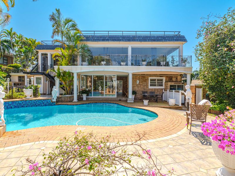 A spacious rear exterior featuring a swimming pool, paved deck, and a two-story residence with a large balcony and stone-clad facade.