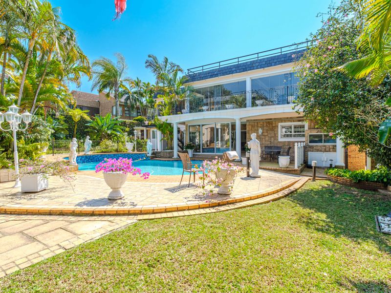 A well-maintained outdoor pool area featuring a paved patio, stone-clad exterior walls, and a spacious upper-level balcony overlooking the garden.