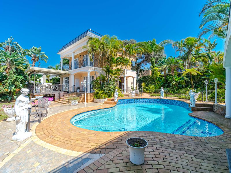 A well-maintained outdoor pool area featuring brick paving, a covered patio, and a balcony overlooking the landscaped garden and swimming pool.