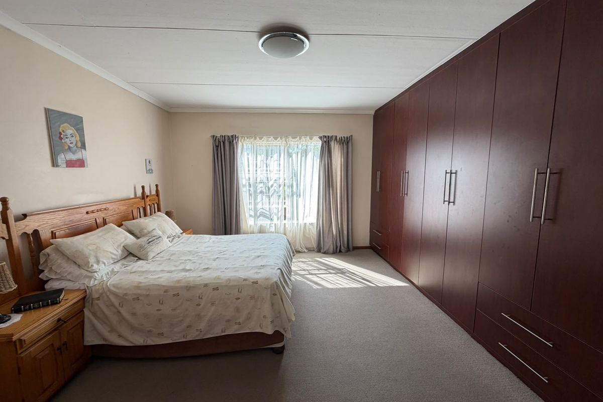 Spacious bedroom featuring extensive wall-to-wall built-in wooden cupboards, neutral carpeting, and natural light through a window with curtains.