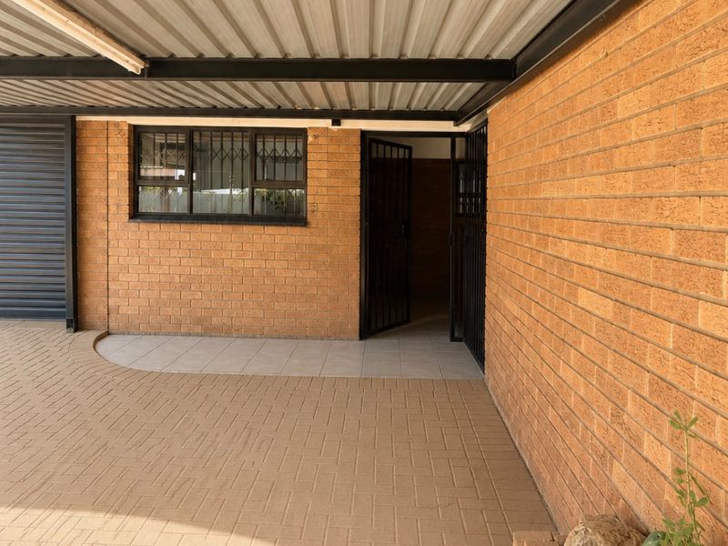 A covered carport area featuring exposed steel beams, brick walls, and a paved driveway with a built-in brick planter.