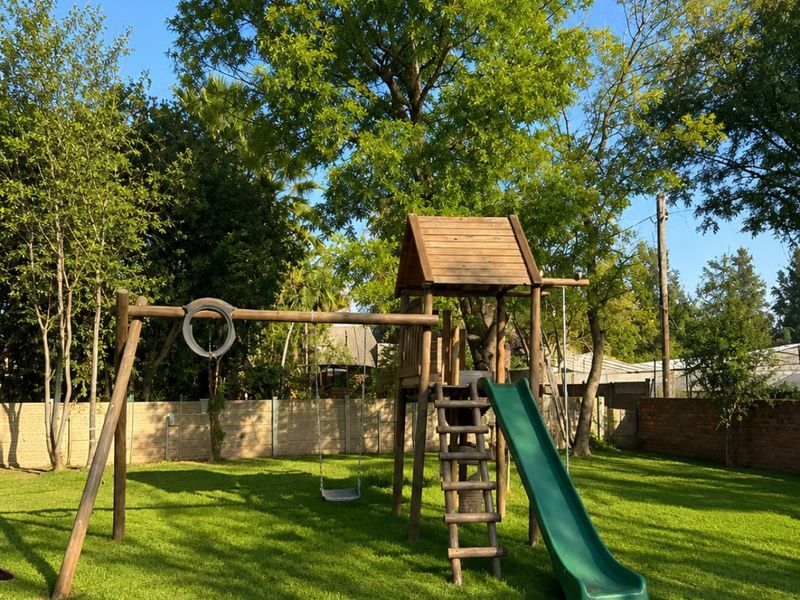 A spacious backyard garden featuring a well-maintained lawn, mature trees, and a wooden playground structure with a slide and swings.