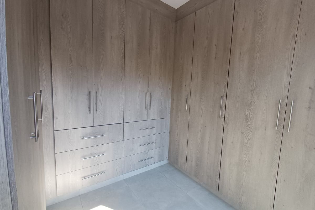 A clean, modern bedroom featuring extensive built-in wooden cabinetry with integrated drawers and sleek metal handles, complemented by neutral floor tiles.