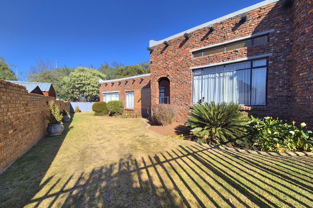 A rear view of the property featuring an exposed brick facade, established garden, and perimeter brick walling under a clear blue sky.
